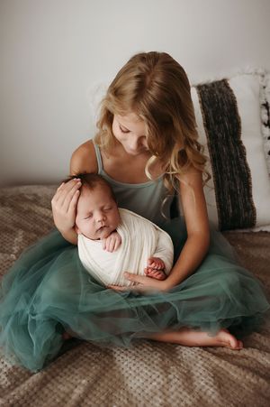 A sister in a teal tulle dress holds her sleeping newborn baby while sitting on a bed with pillows.