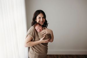 A person in a beige sweater embraces a small newborn baby next to a white window.