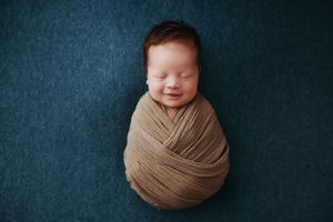 A newborn baby swaddled in beige fabric smiles while sleeping on a dark teal backdrop during a professional photoshoot.