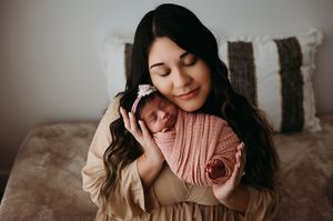 A woman in beige holds a sleeping newborn wrapped in soft pink swaddle on a brown couch.