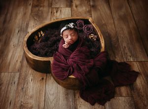 Sleeping newborn wrapped in burgundy blanket nestled in wooden bowl on rustic hardwood floor.