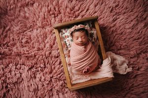 Newborn baby swaddled in pink fabric sleeps peacefully in wooden crate on fluffy pink blanket during photography session.