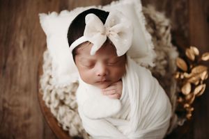 A sleeping newborn swaddled in white fabric with a white bow headband rests in a cozy basket.