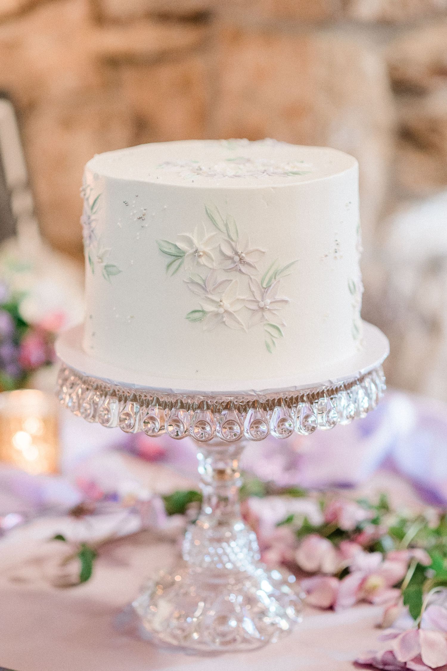 White wedding cake with delicate green leaf details displayed on an elegant crystal cake stand surrounded by pink flowers.