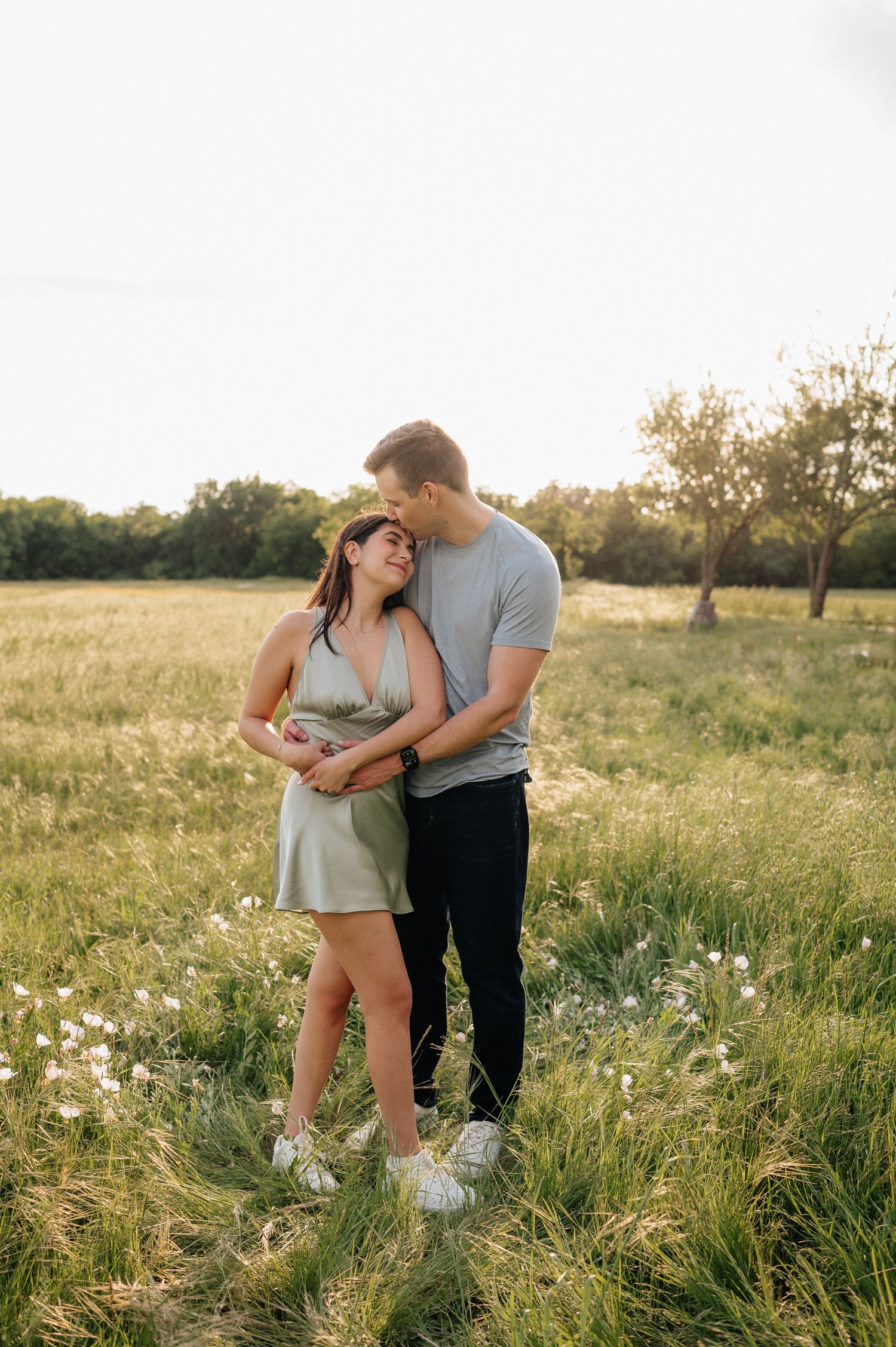 Romantic couple embracing in a sunlit meadow with wildflowers and trees in the golden hour.