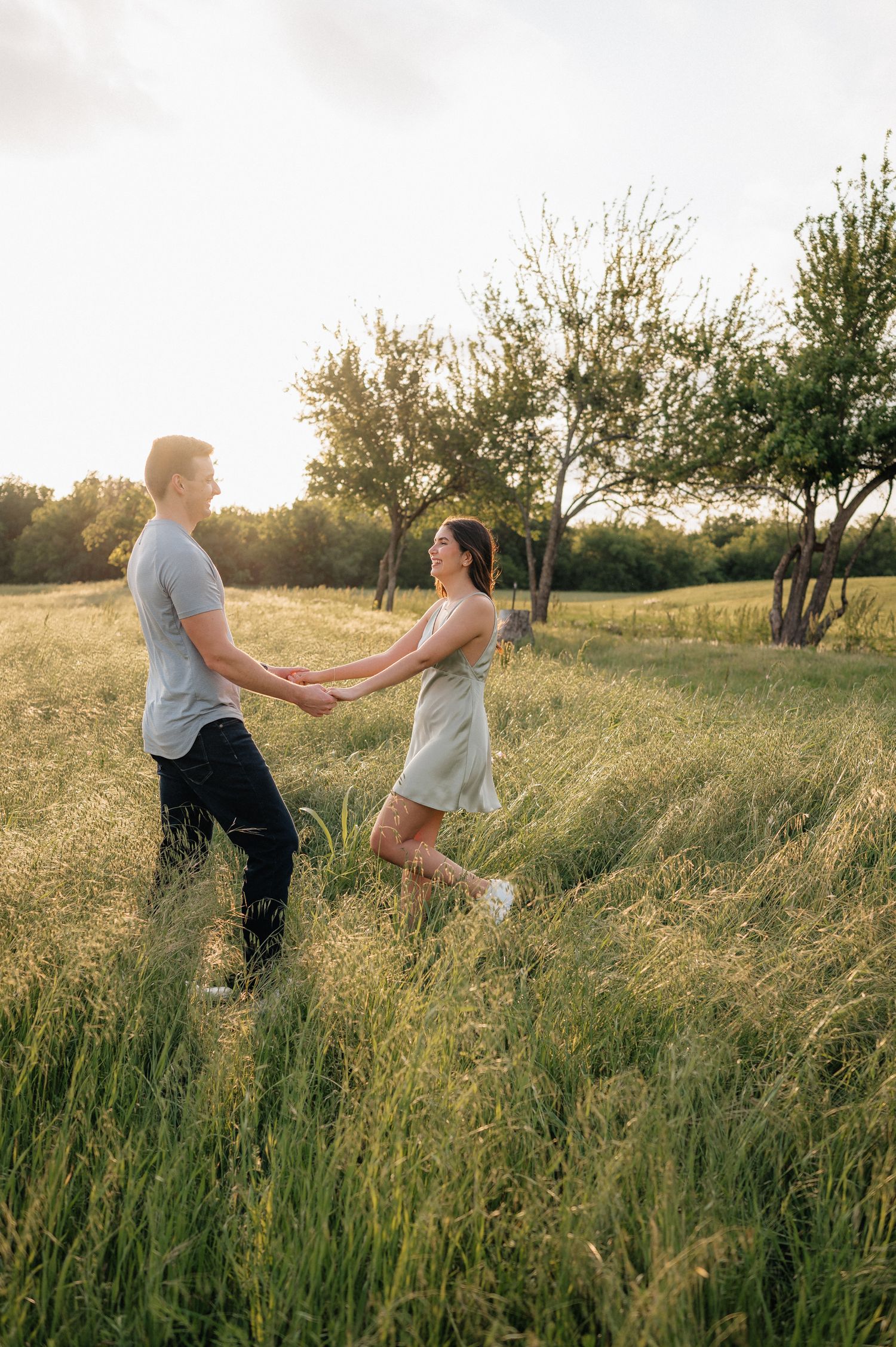 Playful couple dancing together in tall grass with trees in the background during sunset.