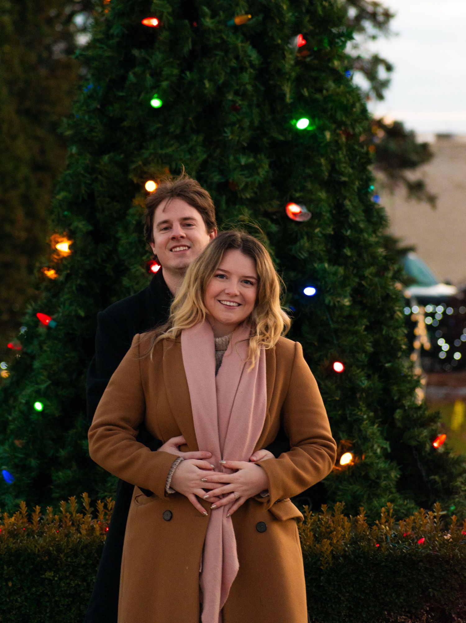 A joyful couple poses in front of an illuminated Christmas tree during evening hours.