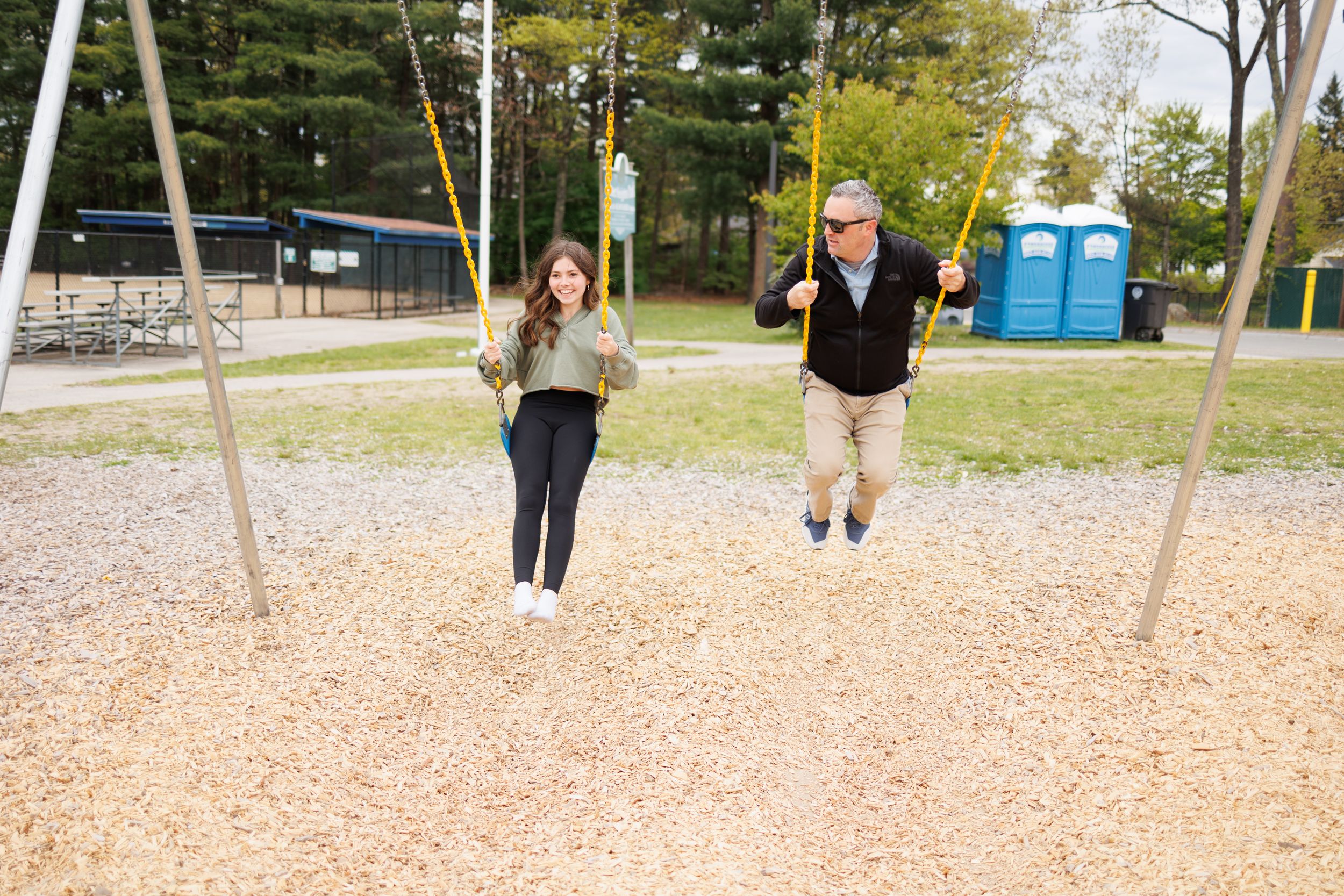 Two people having fun swinging together at a playground with portable restrooms visible in background.