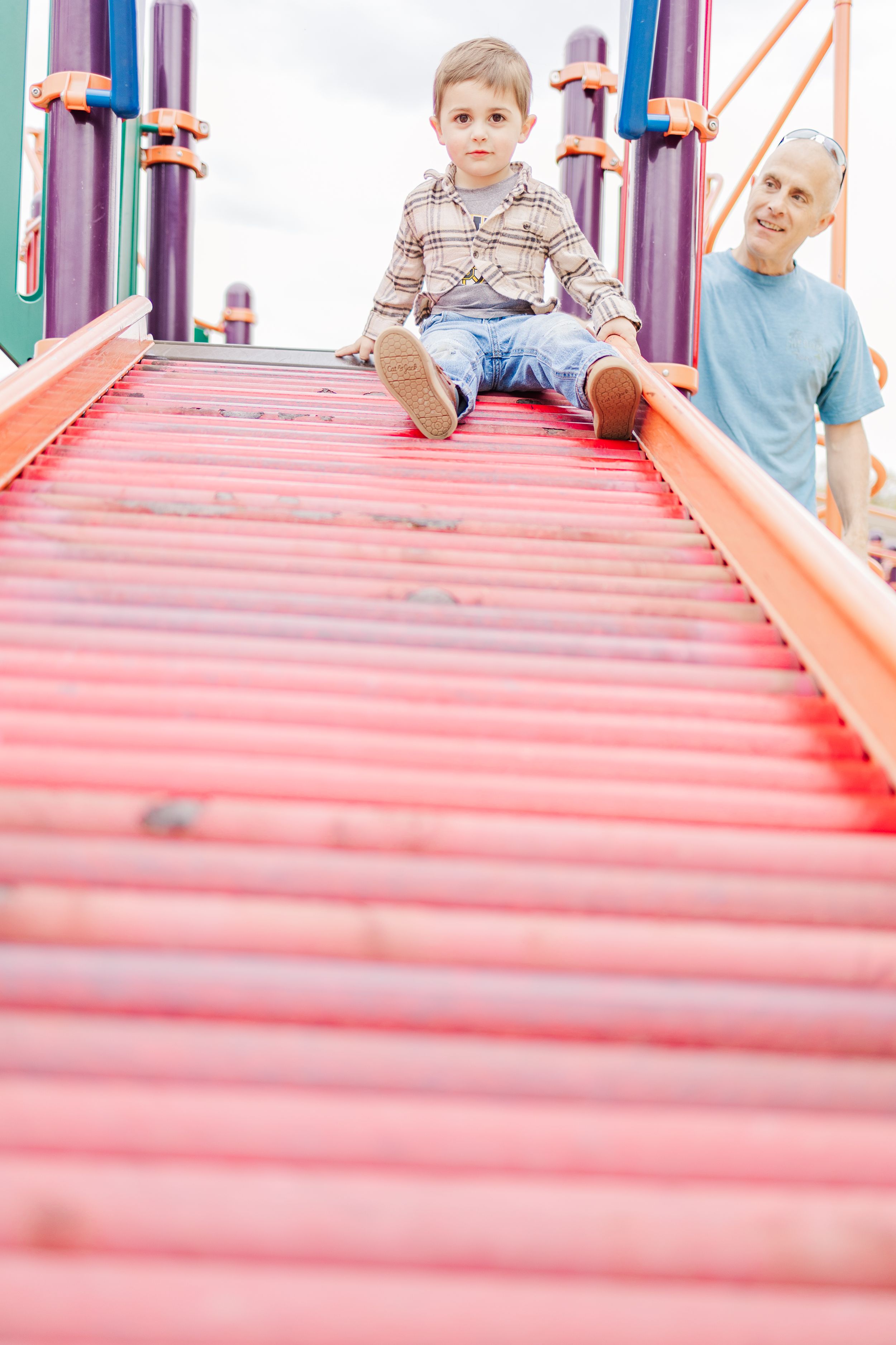 Child sitting on red playground bridge with adult supervising.