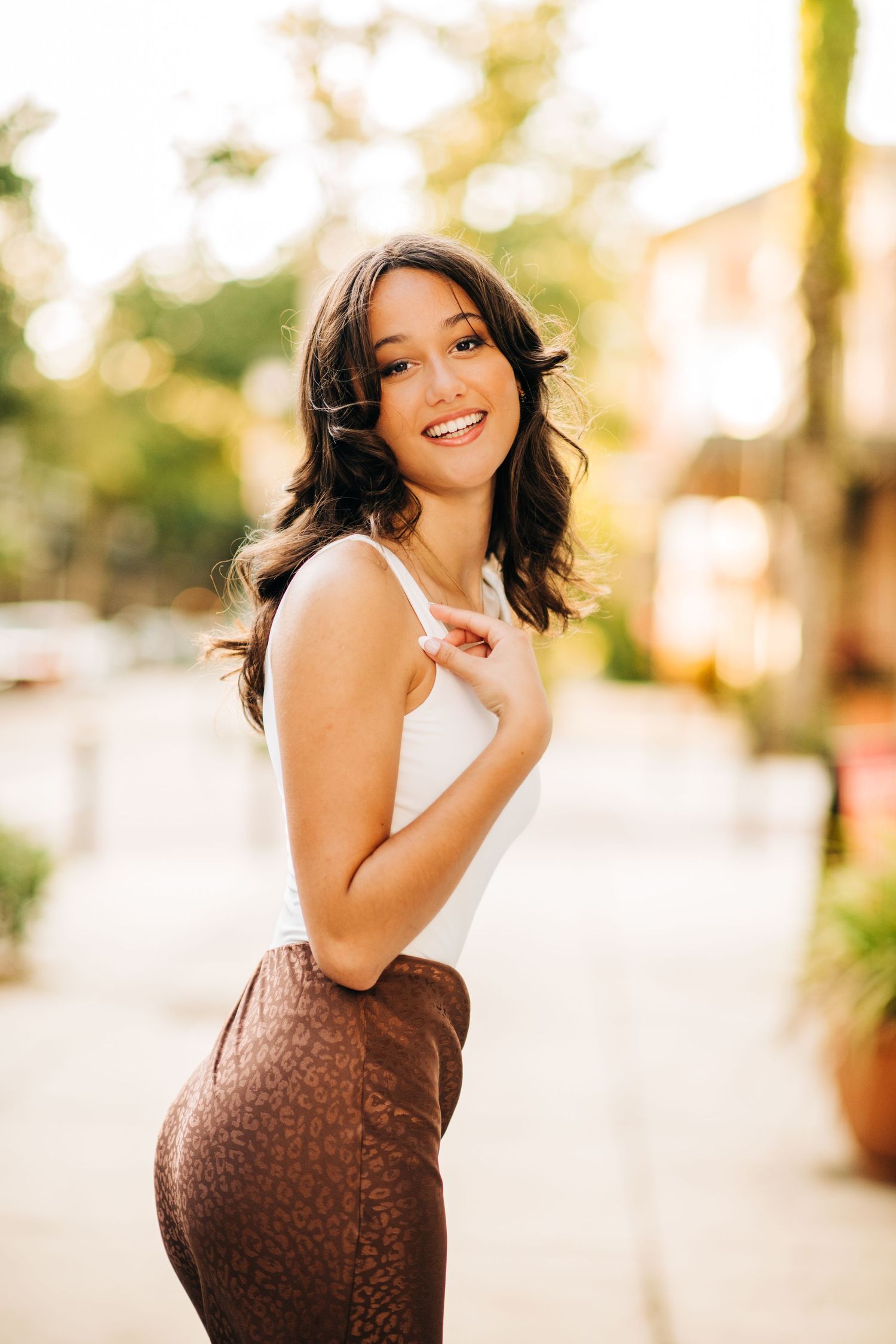 Woman in white top and brown skirt poses outdoors with a cheerful expression in warm sunlight.