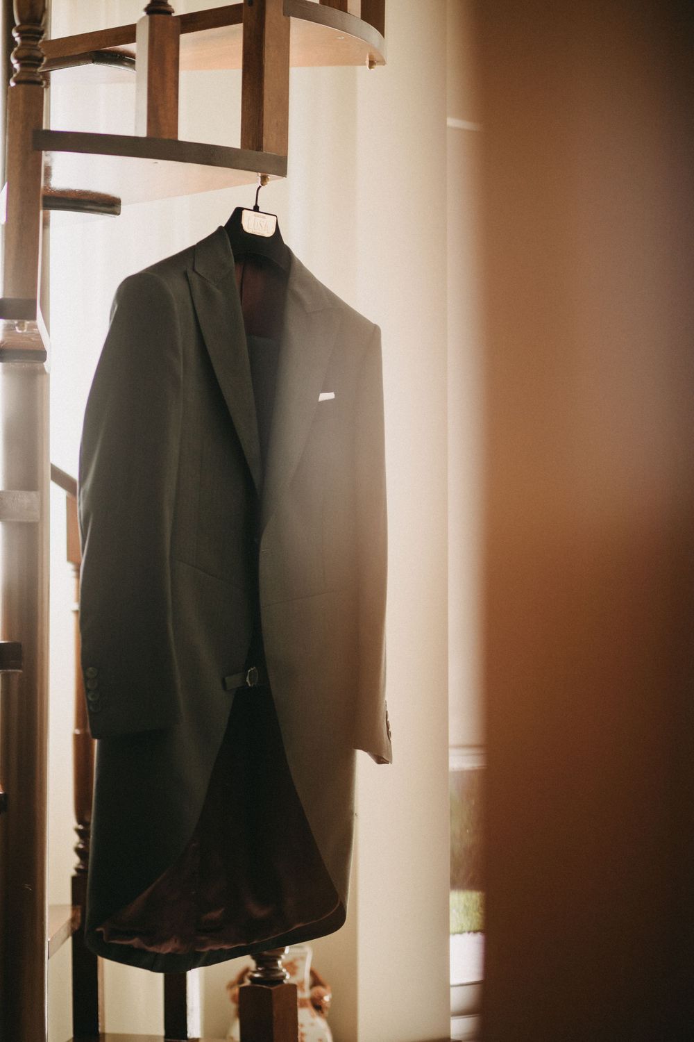 Dark formal suit jacket hanging on wooden rack in soft natural lighting.