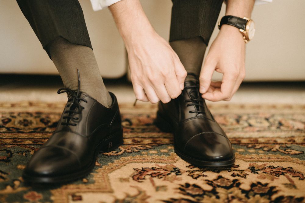 Hands tying black leather dress shoes on ornate patterned carpet.