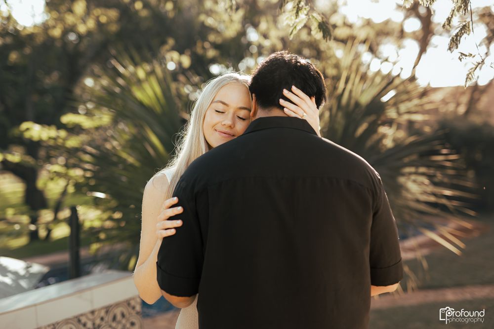 Romantic couple embraces in warm sunlit garden setting with palm trees in background.