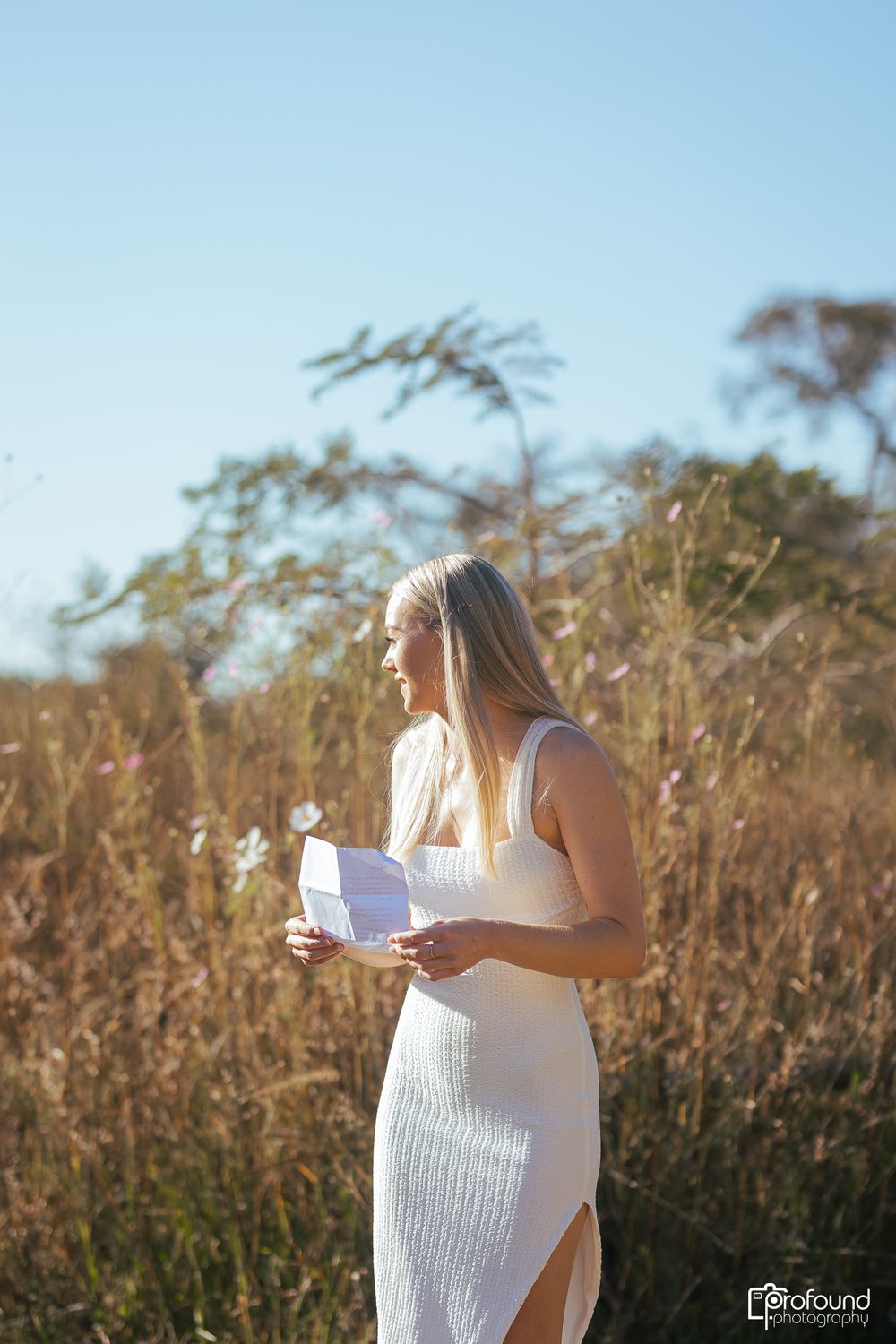 Woman in white dress reading a note in a sunlit field of tall grass.