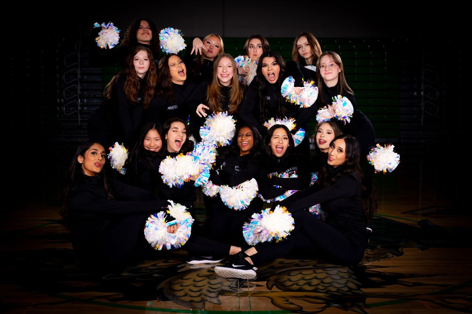 A group wearing black outfits poses with glowing white pom poms against a dark background in a dynamic formation.