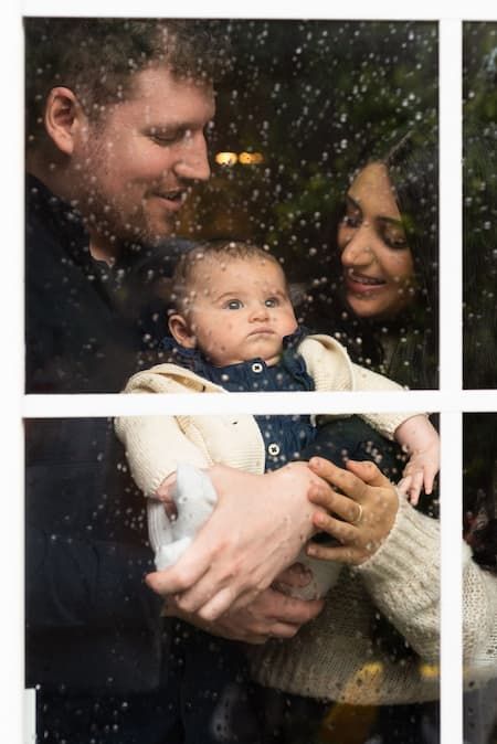Baby looks out the window at the rain with mom and dad as a frame.