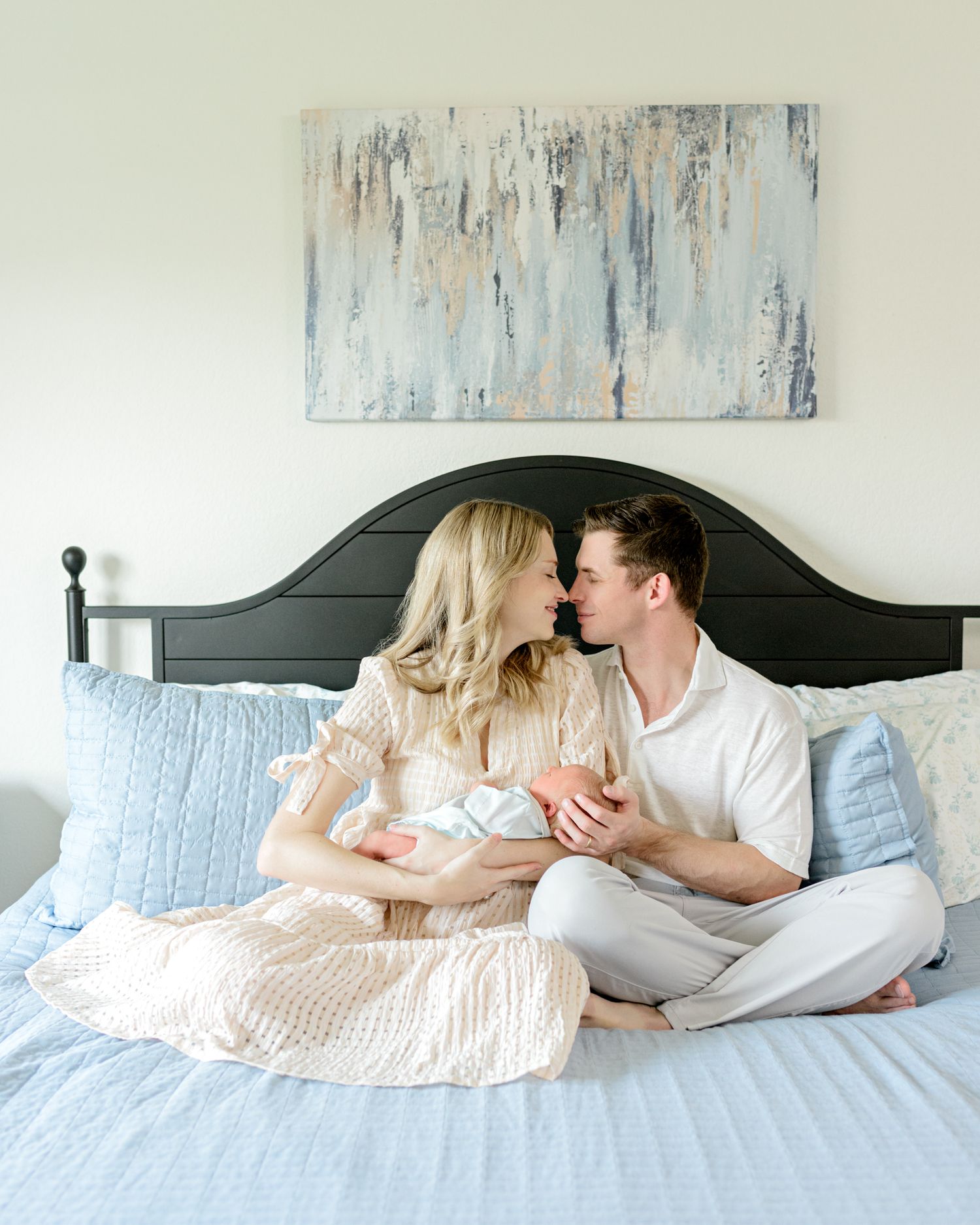 A couple sits on a bed beneath abstract wall art, sharing an intimate moment while holding their newborn baby.