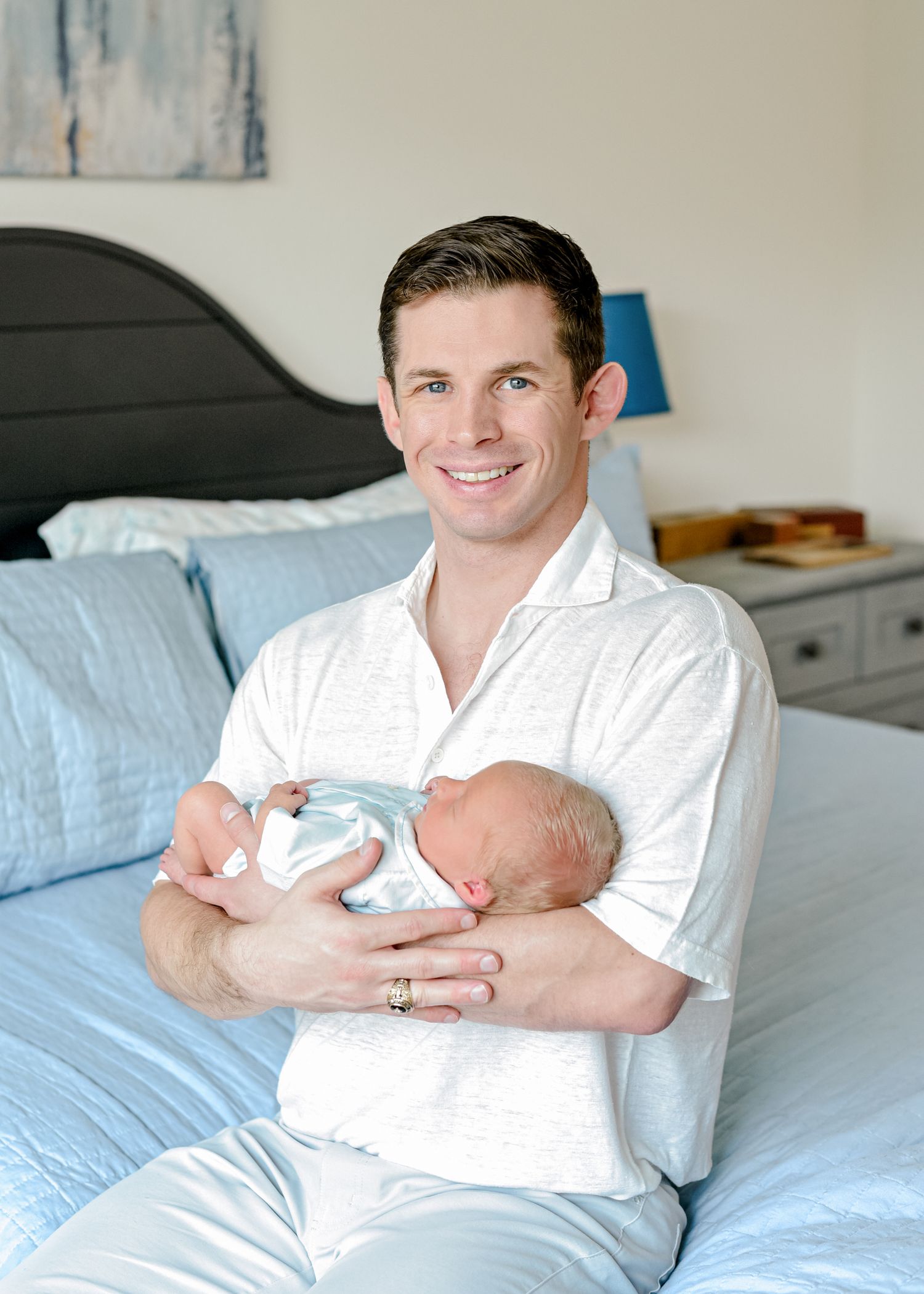 Parent in white shirt holds sleeping newborn against blue bedding beneath modern wall art.