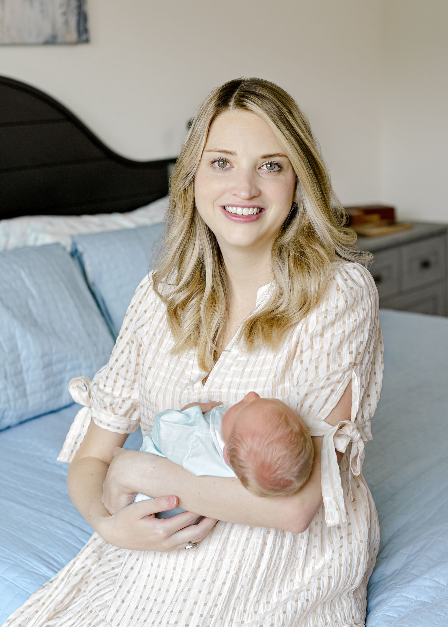 Parent in cream dress holds sleeping newborn while sitting on blue bedding.