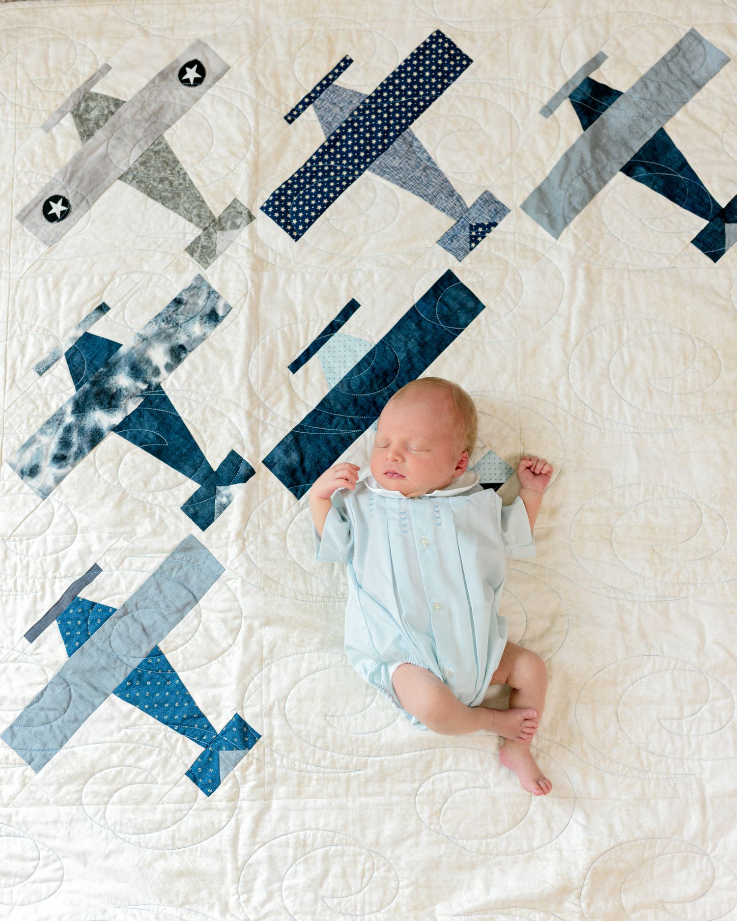 Baby in light blue outfit lying on quilt with patchwork airplane pattern design.