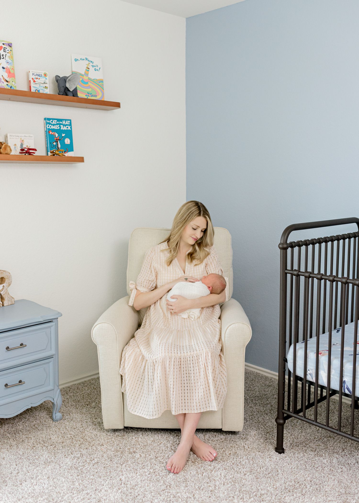 Newborn baby being cradled in a peaceful nursery with wooden shelves and a beige armchair.