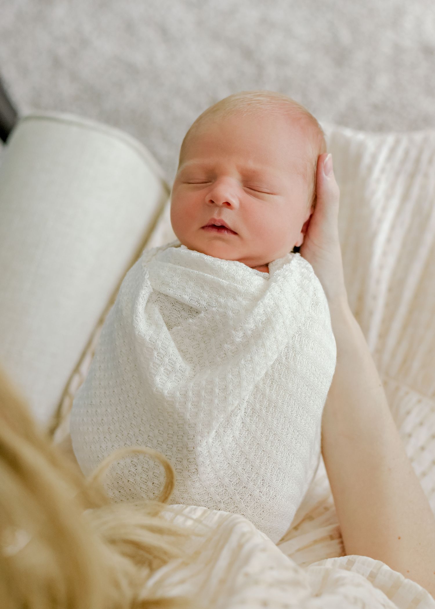 Sleeping newborn wrapped in white knit blanket resting on soft cushion.
