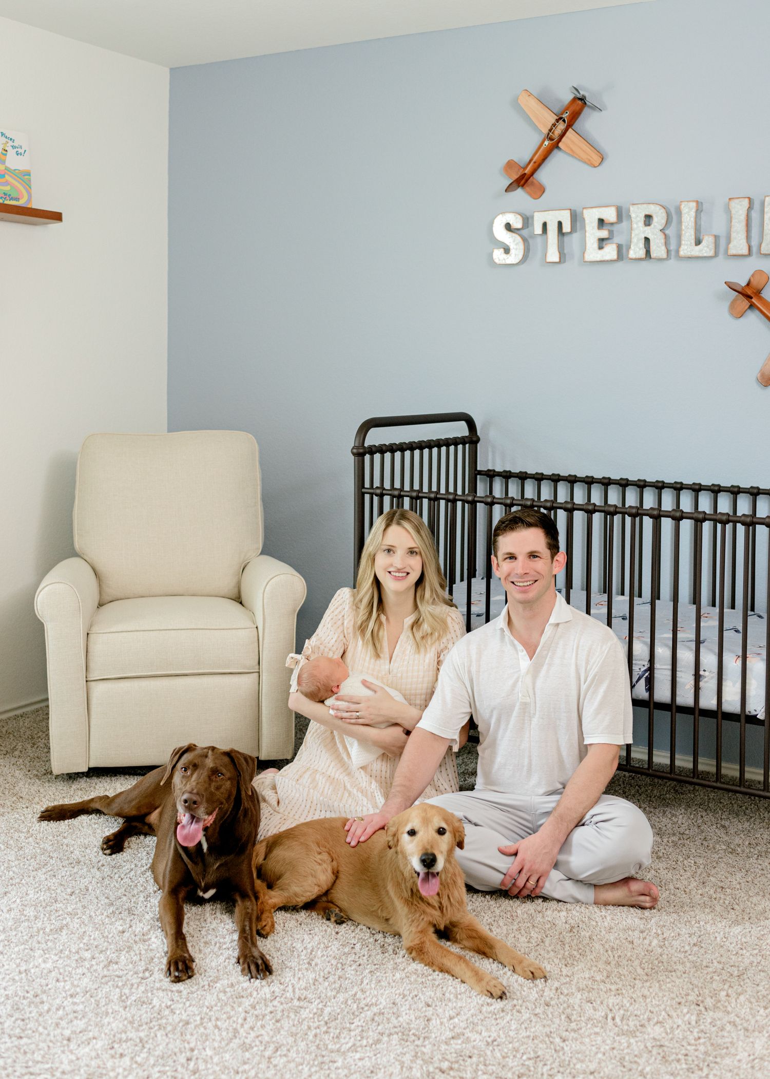 Young couple with newborn and two dogs pose in modern nursery with airplane decor.