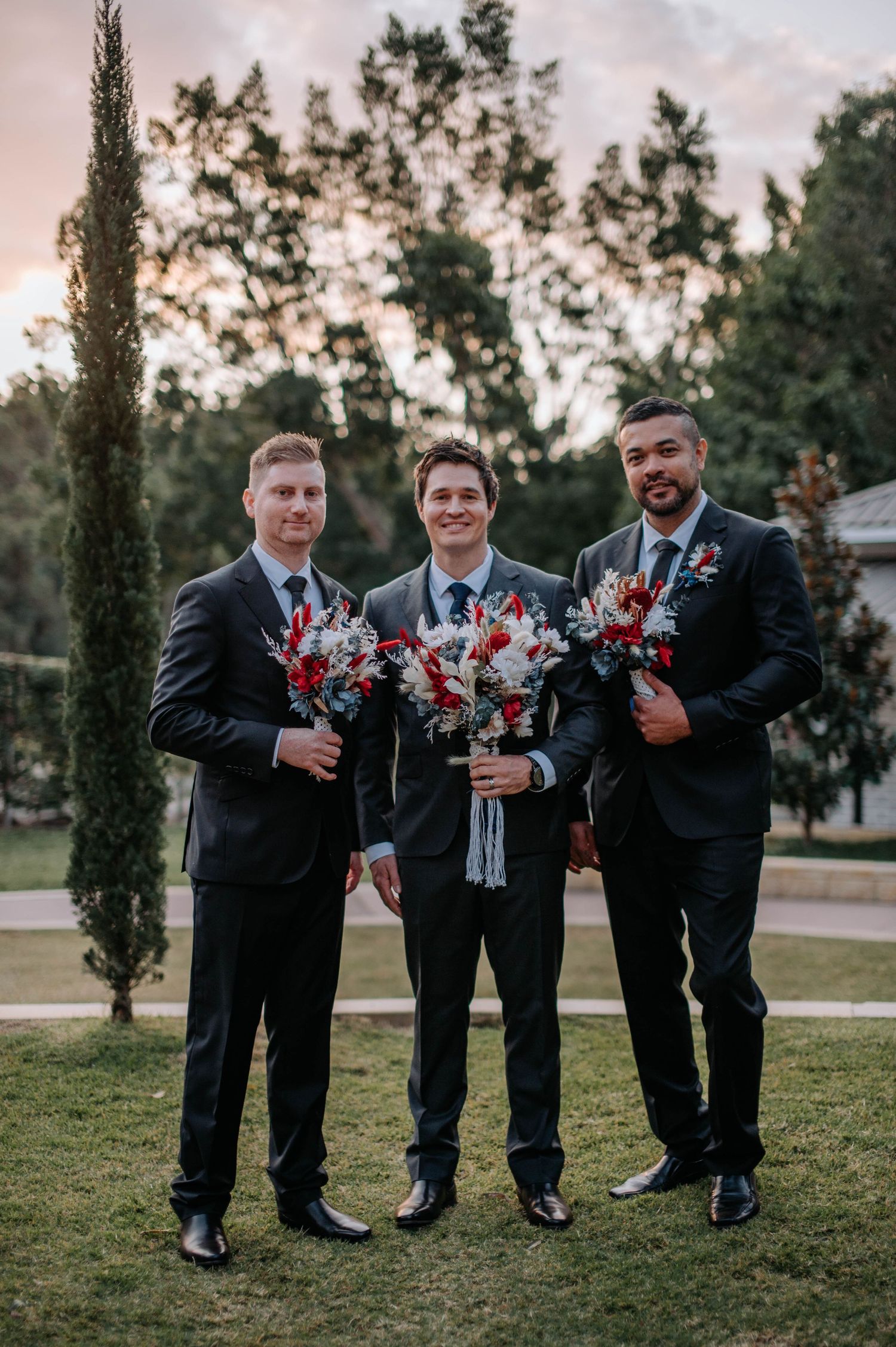 Three groomsmen in formal suits hold colorful bouquets while standing outdoors at dusk.