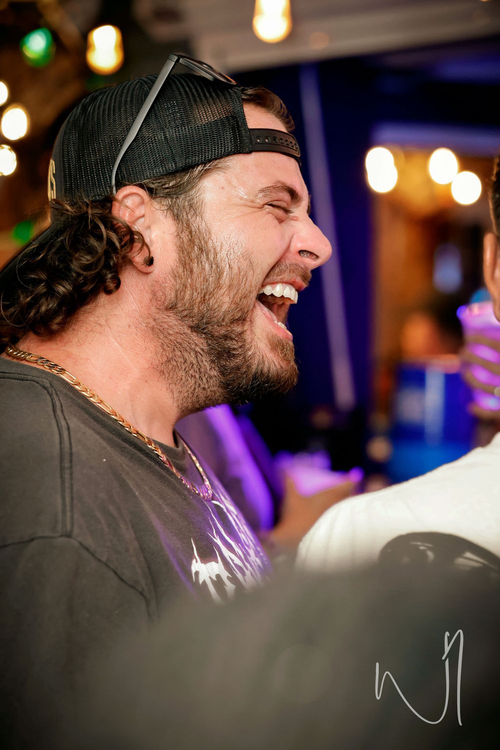 Person wearing a black cap laughing in a dimly lit venue with string lights overhead.