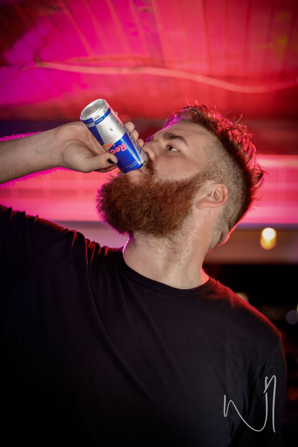 Person with red beard drinking from a can under dramatic red lighting.