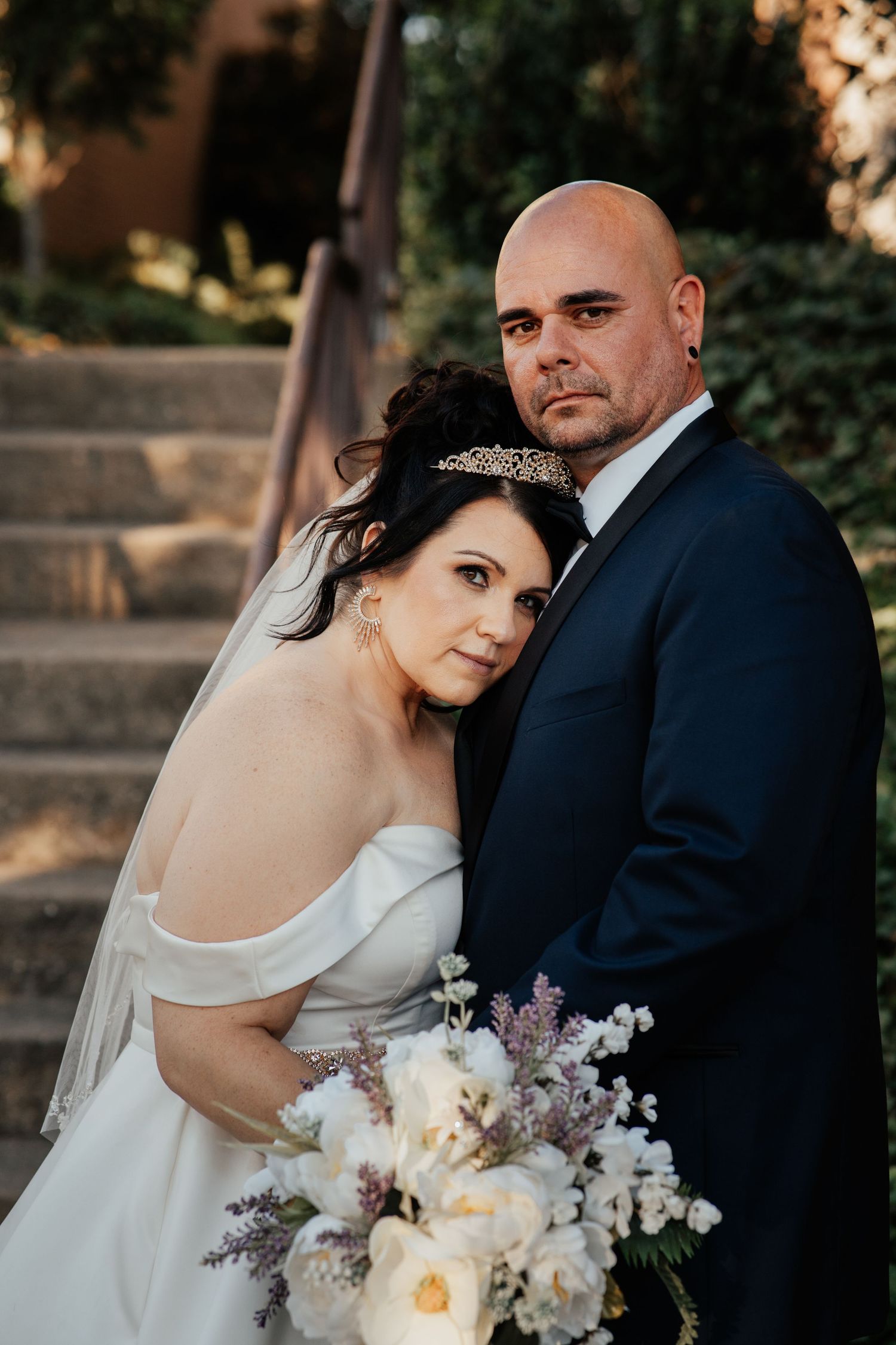 A wedding couple poses on stone steps with an elegant floral bouquet at sunset.