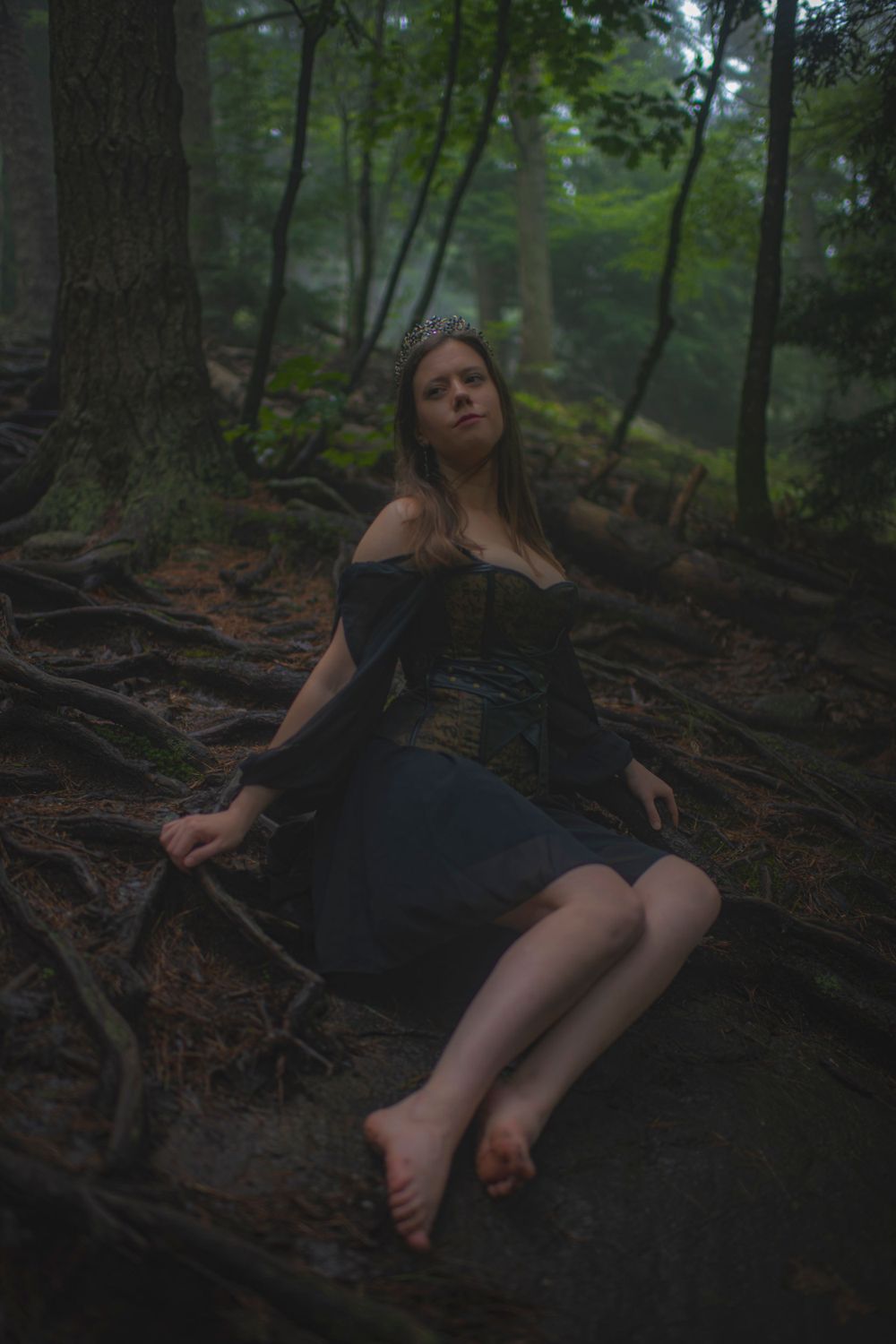 Barefoot figure in dark dress poses dramatically against tree roots in dim forest.