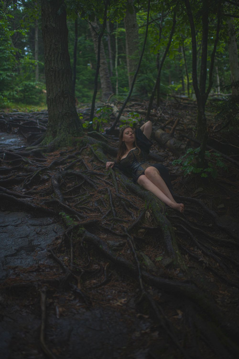 Person in black dress reclining on exposed tree roots by a forest stream.