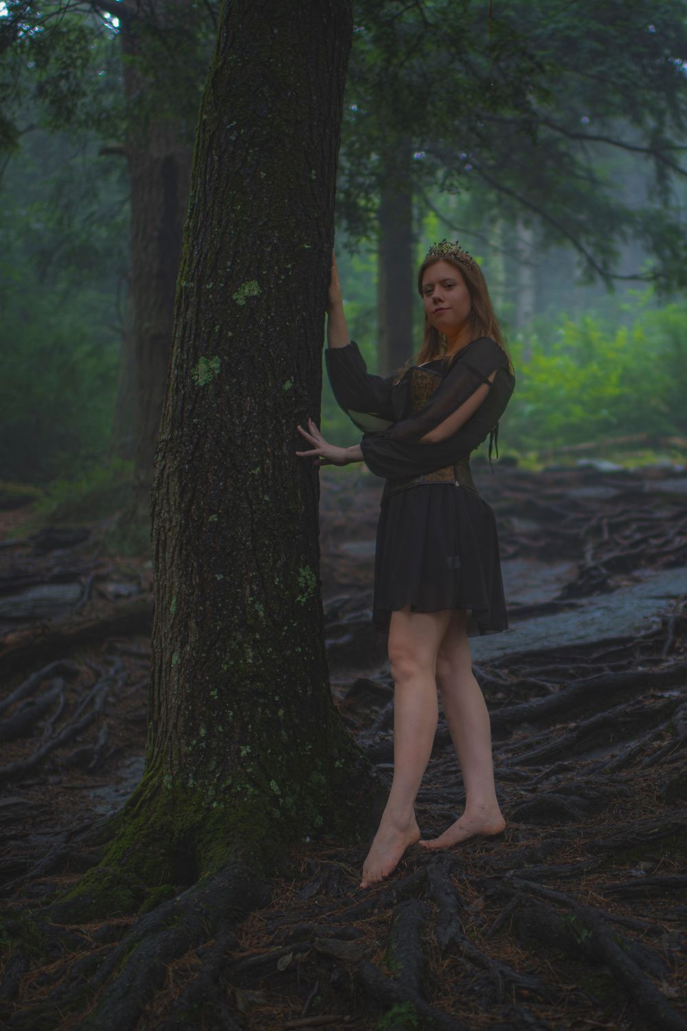 Artistic portrait in dark forest with person in black dress leaning against moss-covered tree.