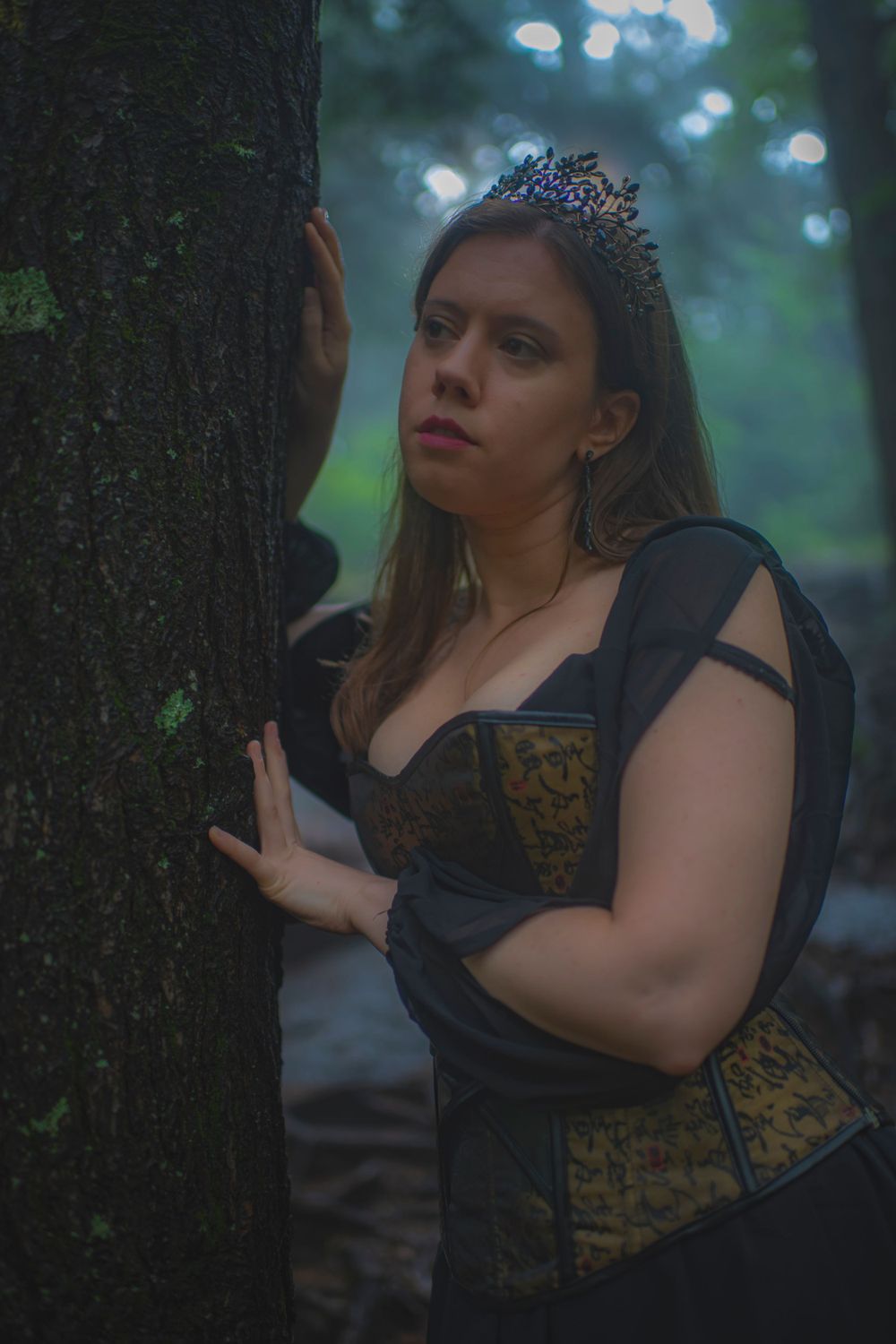 Moody forest portrait with crowned subject in vintage-style corset against tree trunk.