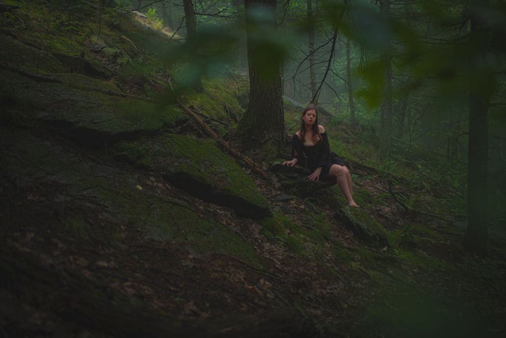 Figure in dark dress sits on mossy forest slope in ethereal lighting.