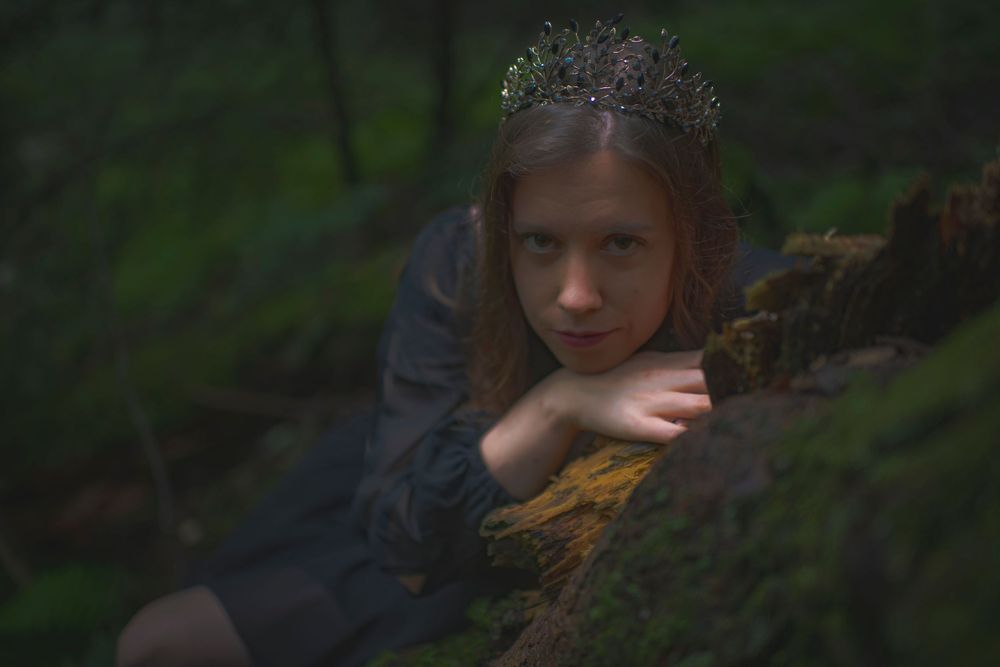 Dramatic portrait in dark forest featuring crowned figure resting on fallen log.