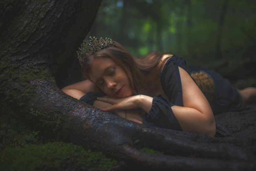 Figure in a dark dress lies dreamily against a tree trunk in a moody forest setting.