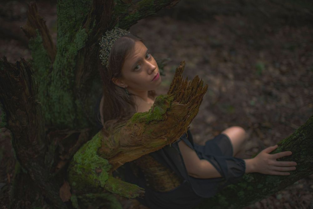 Ethereal forest scene with person in dark dress reaching toward moss-covered tree.