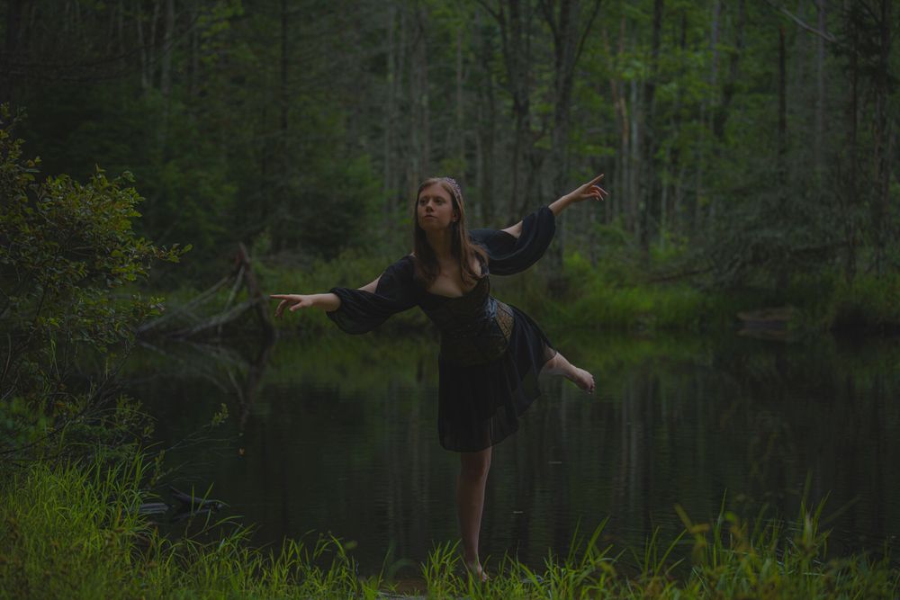 Dancing figure in black dress stands gracefully by a misty forest pond.