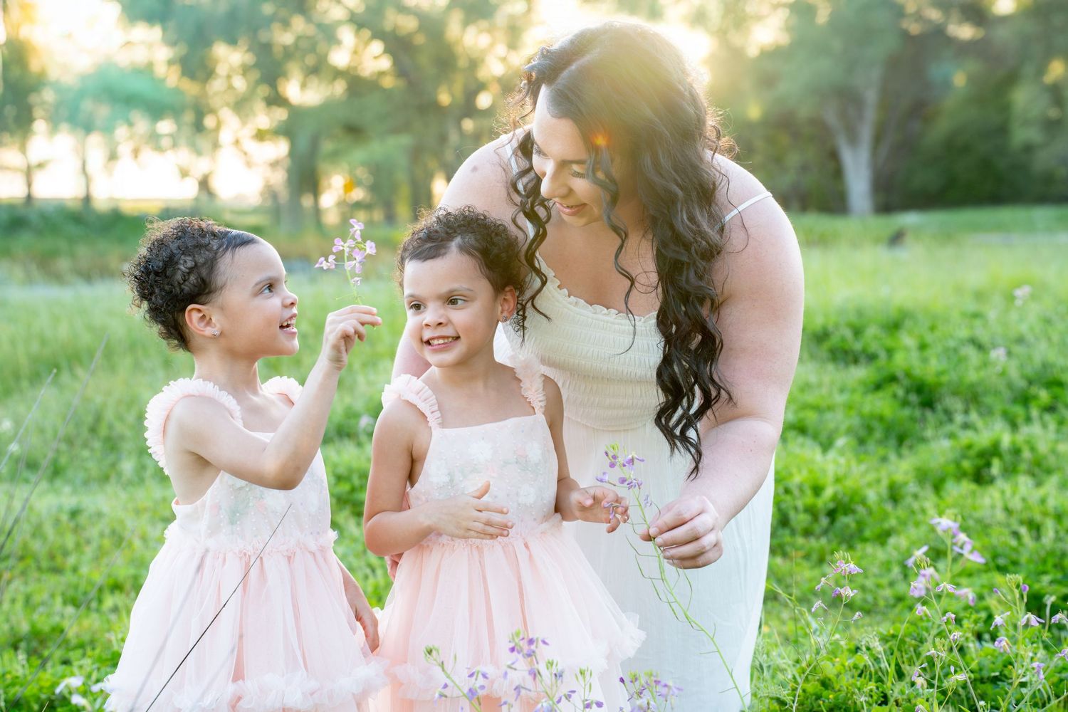 A woman in a white dress stands with two young girls in pink dresses in a grassy field.