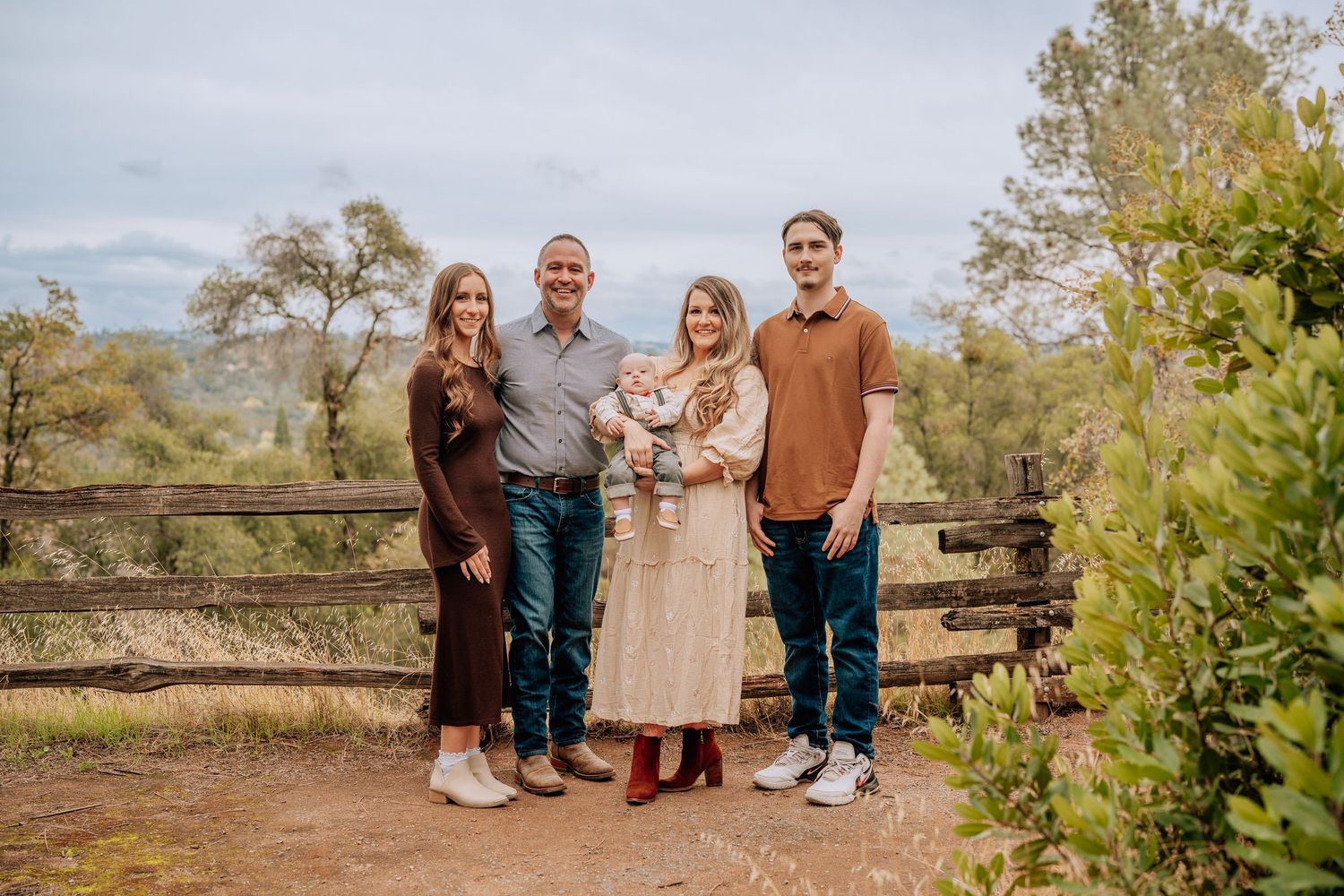 A family poses together in an outdoor setting with rustic wooden fencing and autumn foliage in the background.