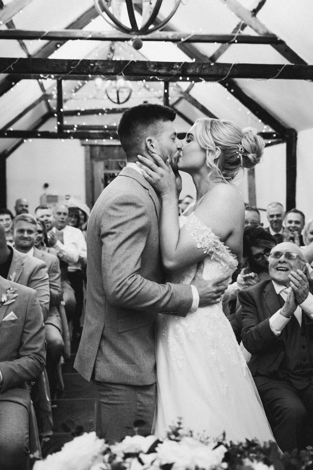 A newlywed couple shares their first kiss as husband and wife in a rustic barn venue filled with their smiling guests.