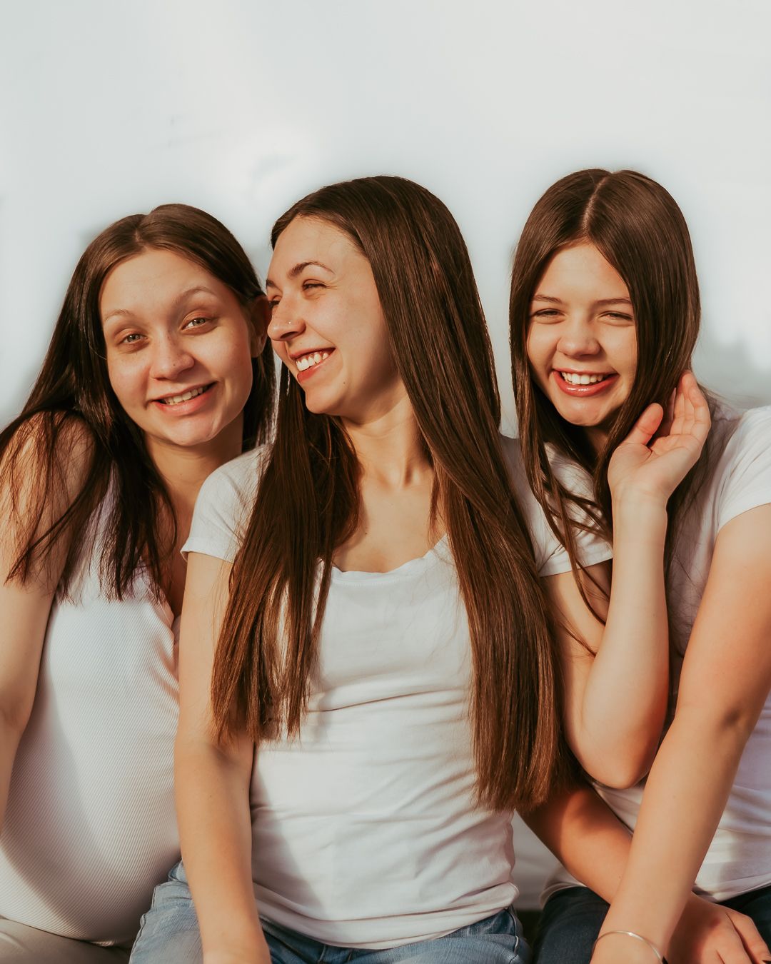 A trio of sisters in white tank tops laugh and smile together in a candid photo.