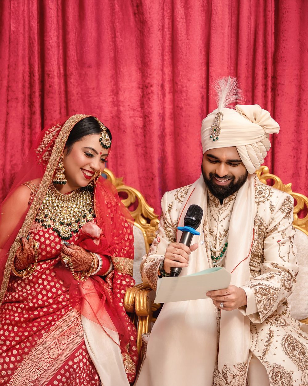 Wedding couple in traditional Indian attire reading messages together against red backdrop.