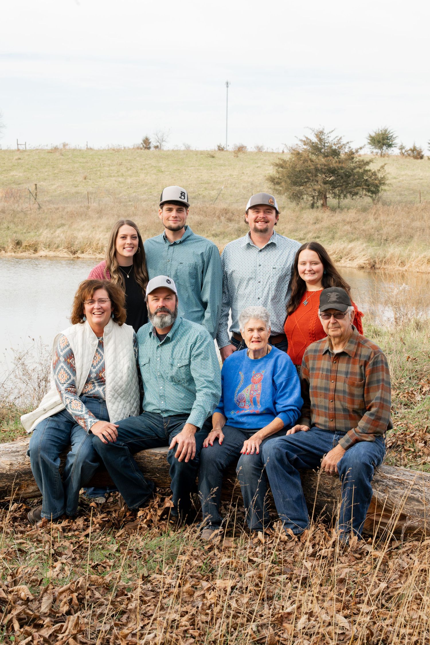 A multi-generational family portrait taken outdoors by a lake with trees and grassland in the background.