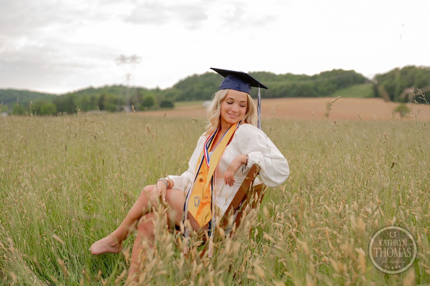 Graduate in academic attire poses on wooden chair in sunlit meadow.