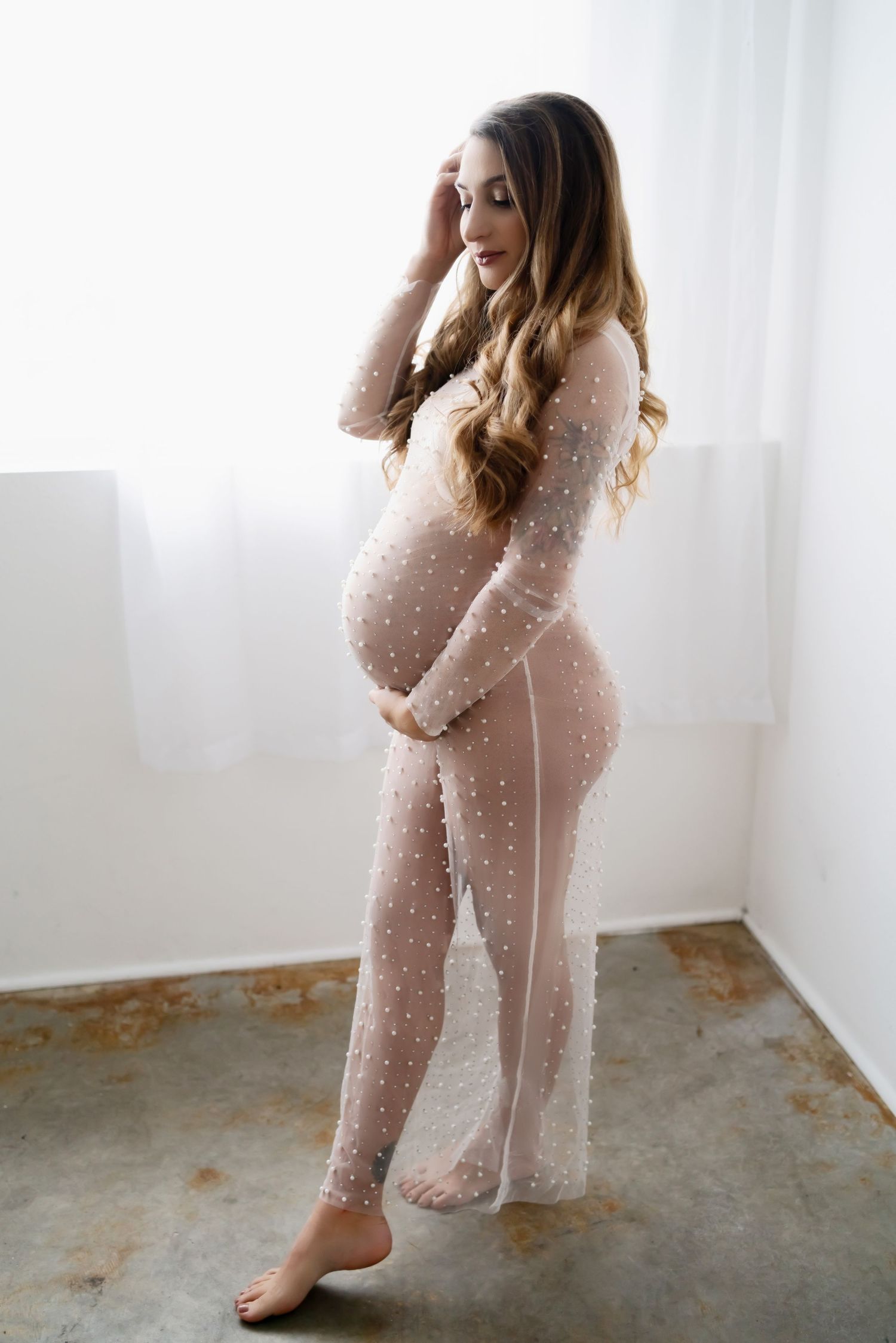 Maternity portrait in sheer beige polka dot jumpsuit against white backdrop.
