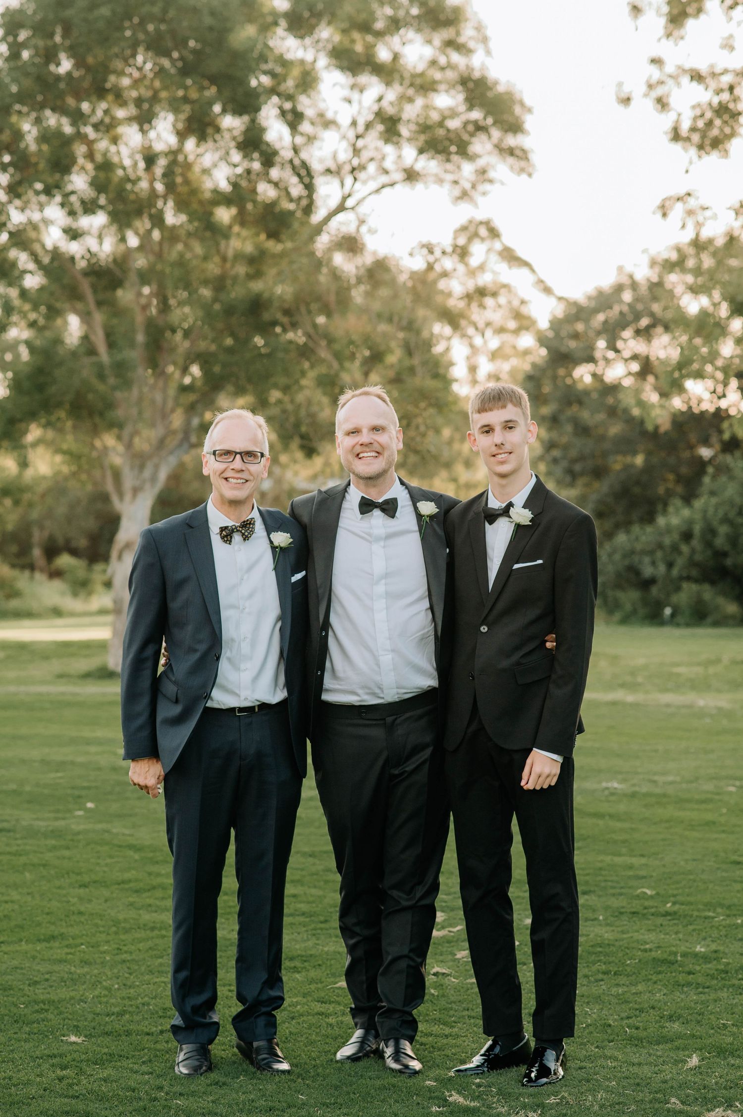 Three groomsmen in black tuxedos and bowties pose together on a green lawn at sunset.