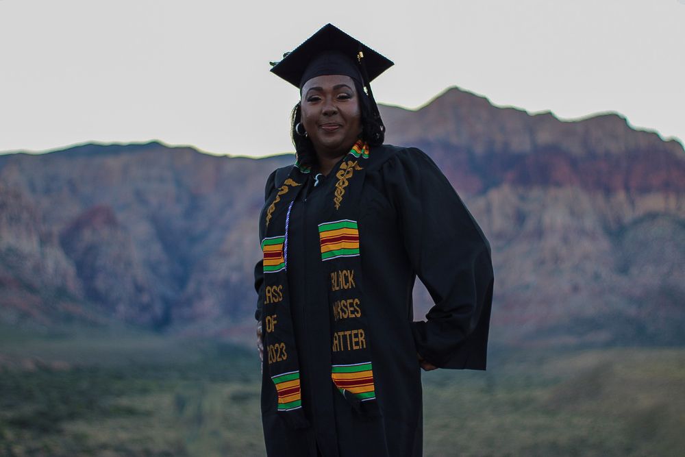 Black woman in graduation cap and gown with Kente cloth stole stands before red rock canyon mountains.